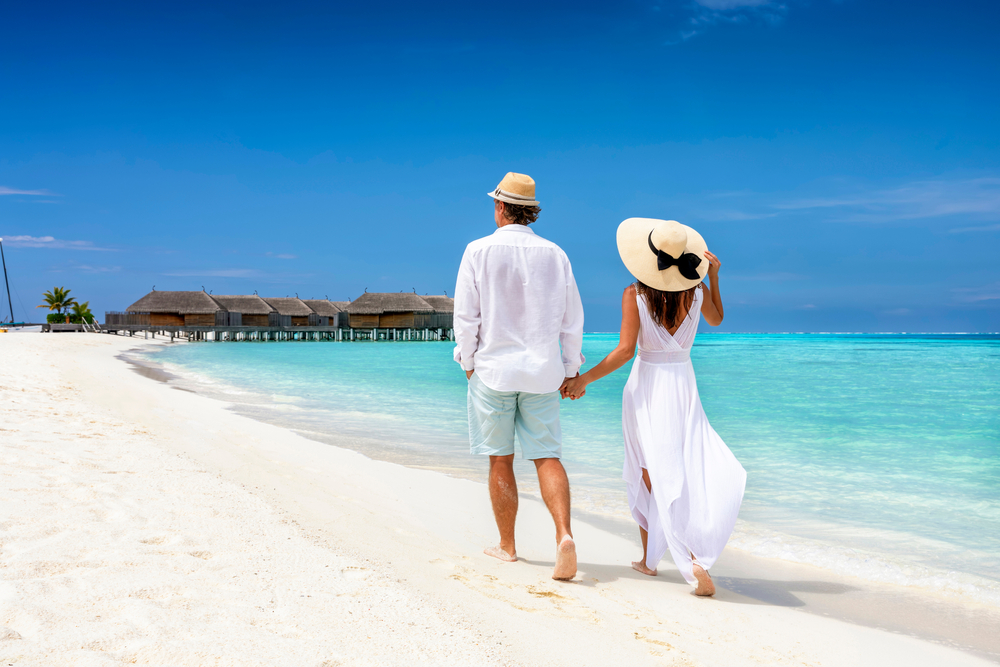 Happy couple in white clothing with hats walks down the beach holding hands