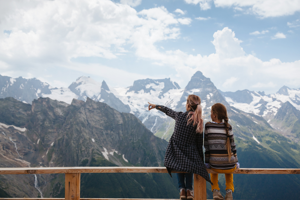 Mom with daughter relaxing in the rustic wooden terrace on mountain, alpine view, snow on hills. Dombay, Karachay-Cherkessia, Caucasus, Russia.