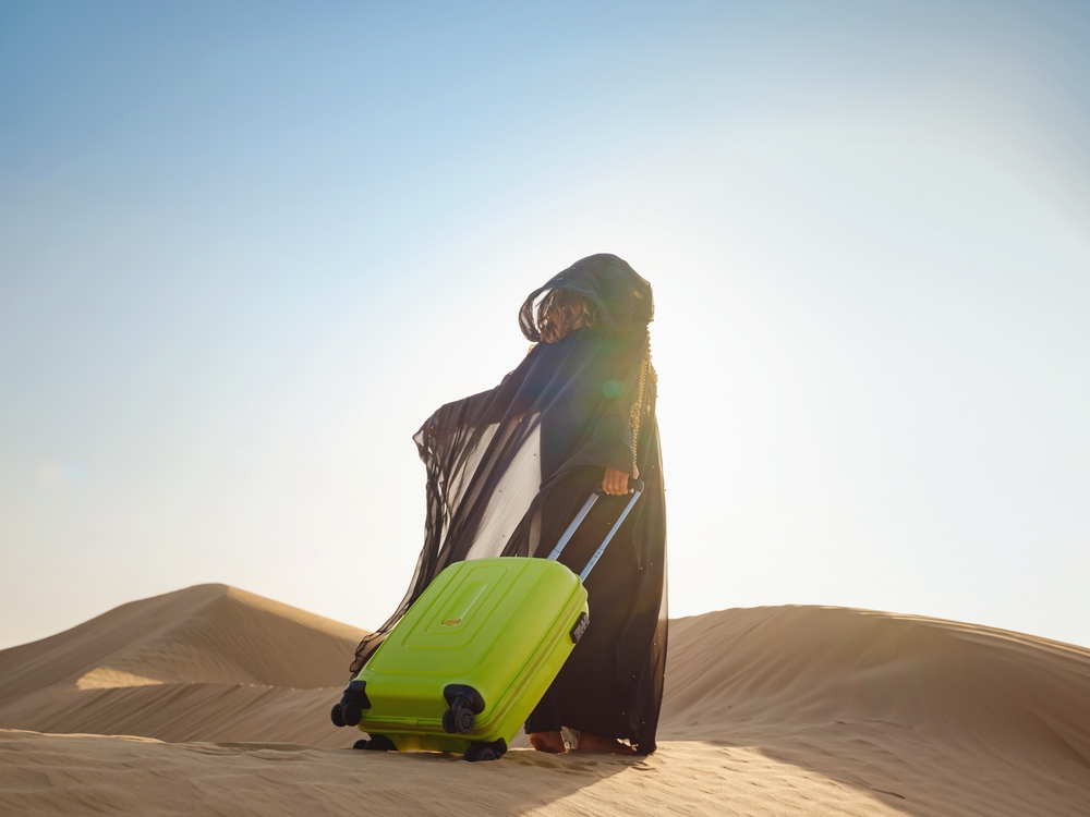 Arabian woman in traditional abaya costume with bright light green or yellow suitcase in sands. Concept and idea of travel to Oman
