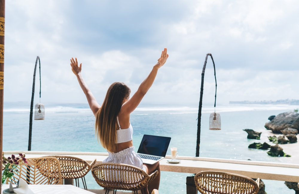 Back view of anonymous female freelancer sitting at table on wicker chair with raised arms and enjoying picturesque seascape during vacation in Bali