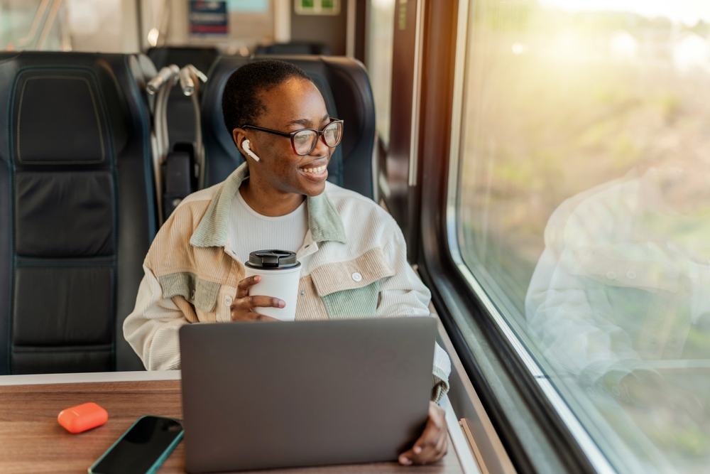 Happy African young businesswoman enjoying in train ride while drinking takeaway coffee. Business trips, tourist solo travel.