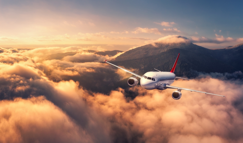 Airplane is flying above mountain peak in orange low clouds at sunrise. Landscape with passenger airplane, red sky, hills in fog. Aircraft is taking off. Business travel. Commercial plane. Aerial view
