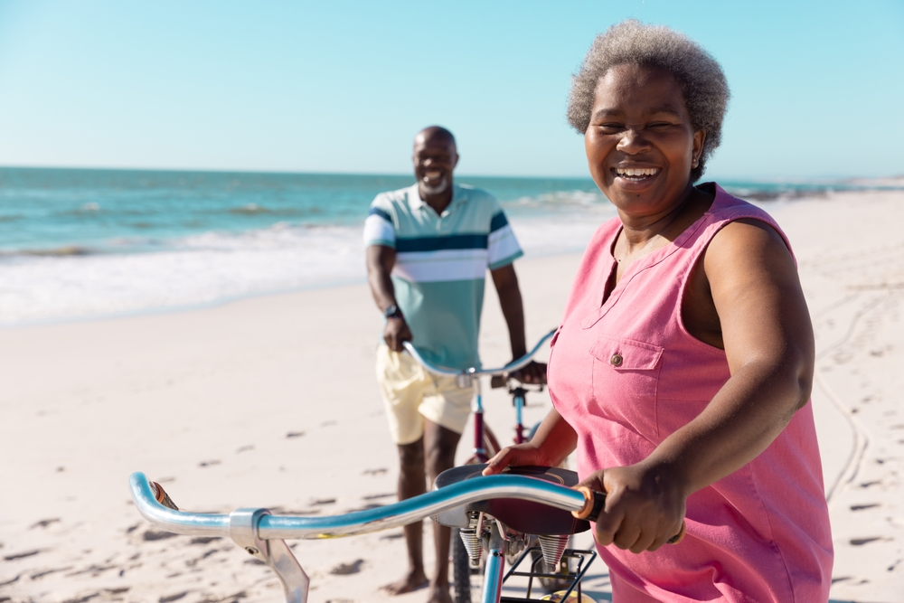 Smiling african american senior couple with bikes walking on sandy beach under clear sky in summer.
