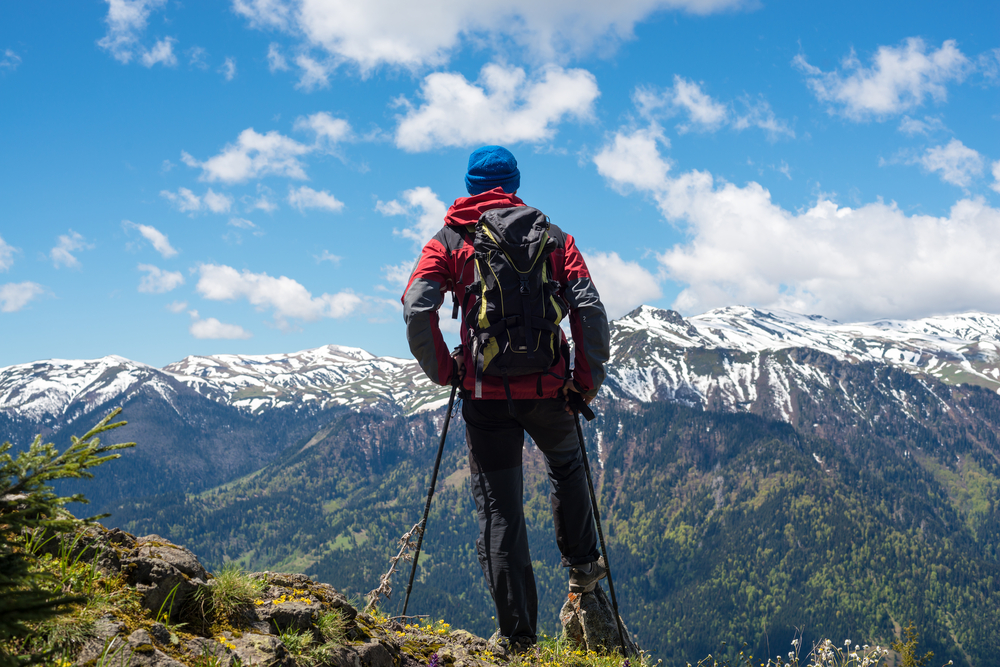 Adventurer, with backpack, stands on the mountain top and admires snow covered mountain ranges stretching to horizon in blue haze. Epic travel in the mountains. Back view. 