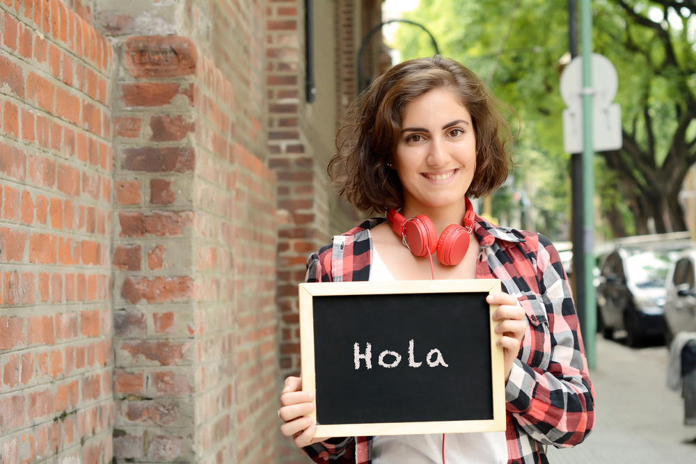 Young beautiful woman holding chalkboard with text "Hola". Outdoors.