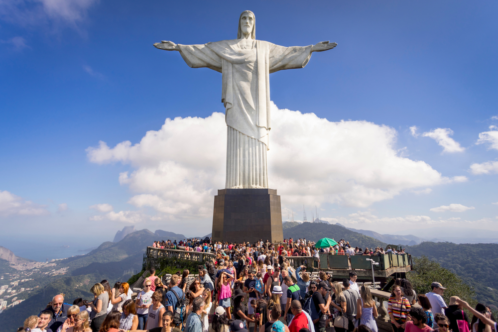 Clear view of a crowd of tourists on top of the Corcovado mountain in Rio de Janeiro with the Christ statue towering over them and a cloud behind in a blue sky