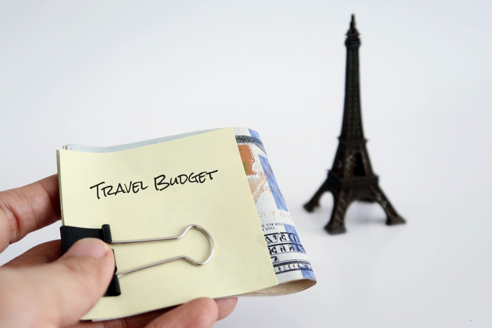 Hand holding money clip with a note TRAVEL BUDGET - with blurred miniature Paris Eiffle Tower on background, concept of budget saving for trip of a lifetime,holiday travel fund planning