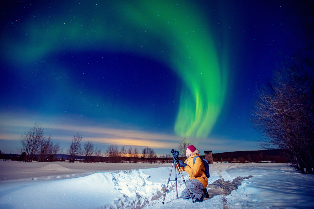Concept photo tour to arctic travel, photographer man with camera and tripod photographs aurora borealis, northern lights green.