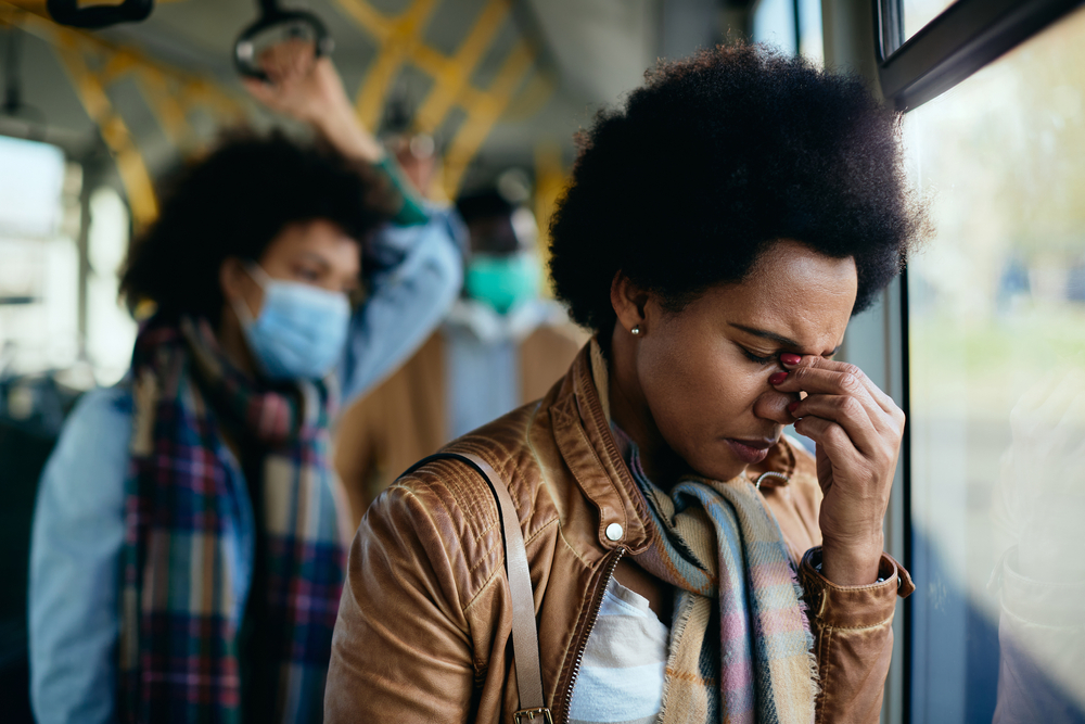 Displeased black woman holding her head in pain while traveling by public transport without protective face mask during COVID-19 pandemic.