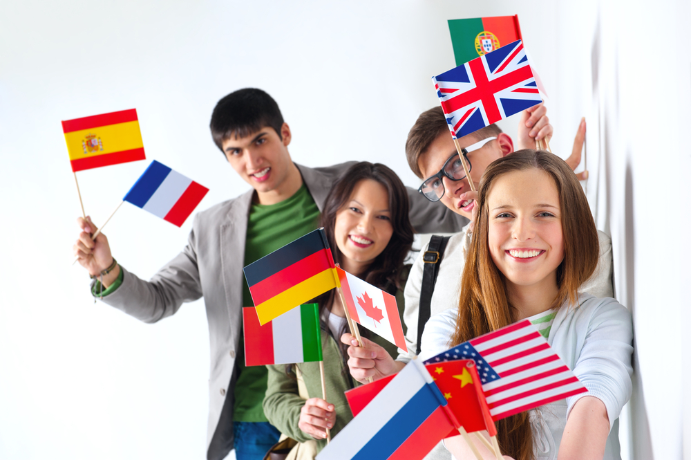 Group of cheerful happy students holding international flags and looking at camera leaning on white wall at campus. International education concept