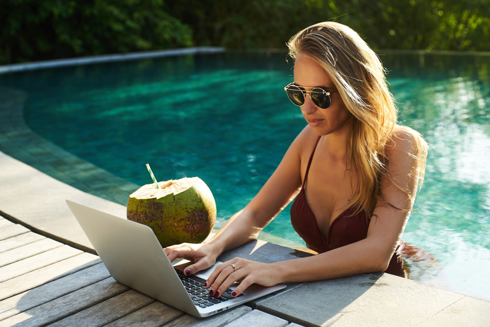 Attractive female wearing sunglasses and swimsuit sitting in the swimming pool and working on computer