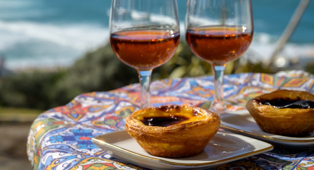 Portugal's traditional food and drink, glasses of porto wine or muscatel de setubal, sweet dessert Pastel de nata egg custard tart pastry served with view on blue Atlantic ocean near Sintra in Lisbon