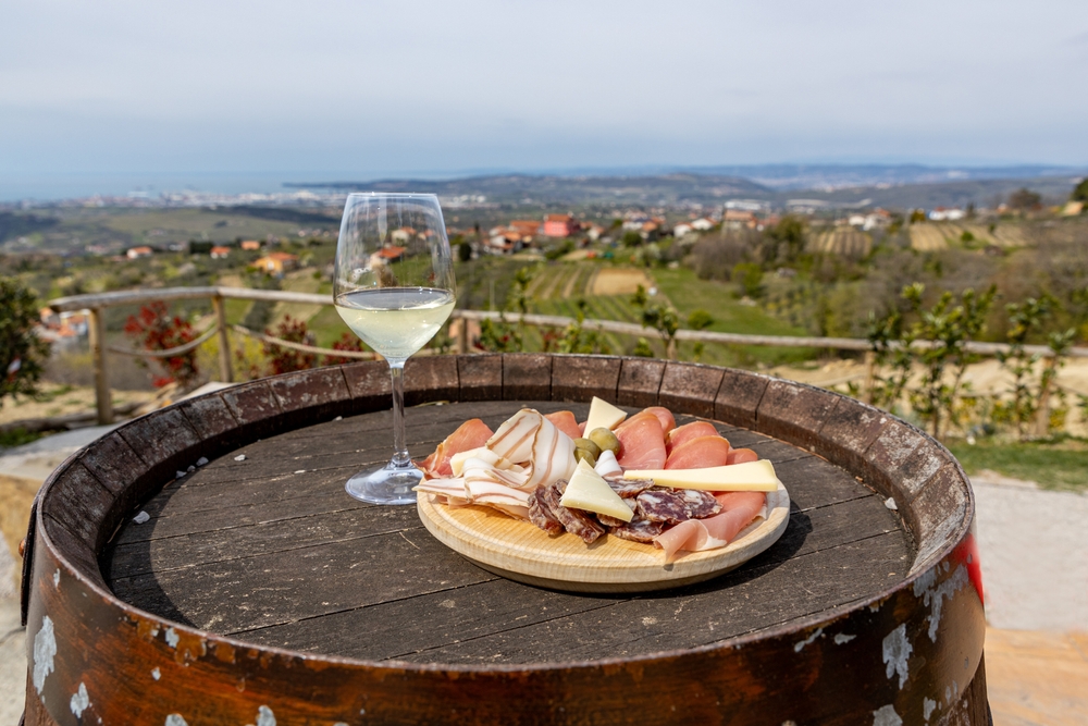 A glass of wine and prosciutto, cheese and olive tapas on a wine barrel overlooking the green fields and the Adriatic