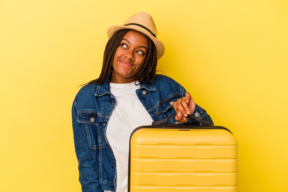 Young african american traveler woman holding suitcase isolated on yellow background dreaming of achieving goals and purposes
