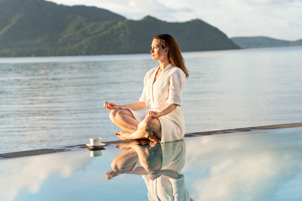 brunette in white robe sits at edge of pool, holding coffee mug and meditating during sunrise.