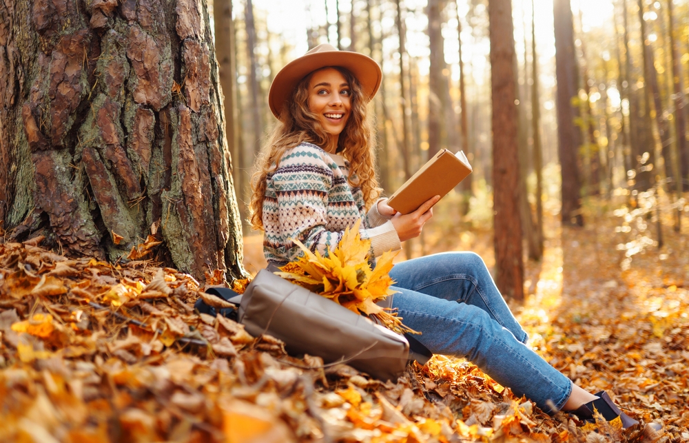 A tourist female in a hat sits in the autumn forest on yellow leaves and reads a book. Beautiful woman enjoying sunny autumn weather.