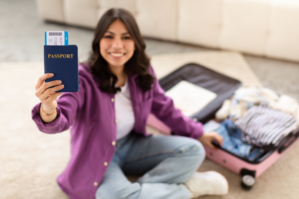 A smiling young middle eastern woman sits on the floor, extending a passport towards the camera with a suitcase beside her