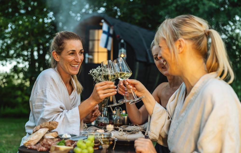 People in bathrobes toasting with wine glasses near a Finnish mobile sauna barrel outdoors