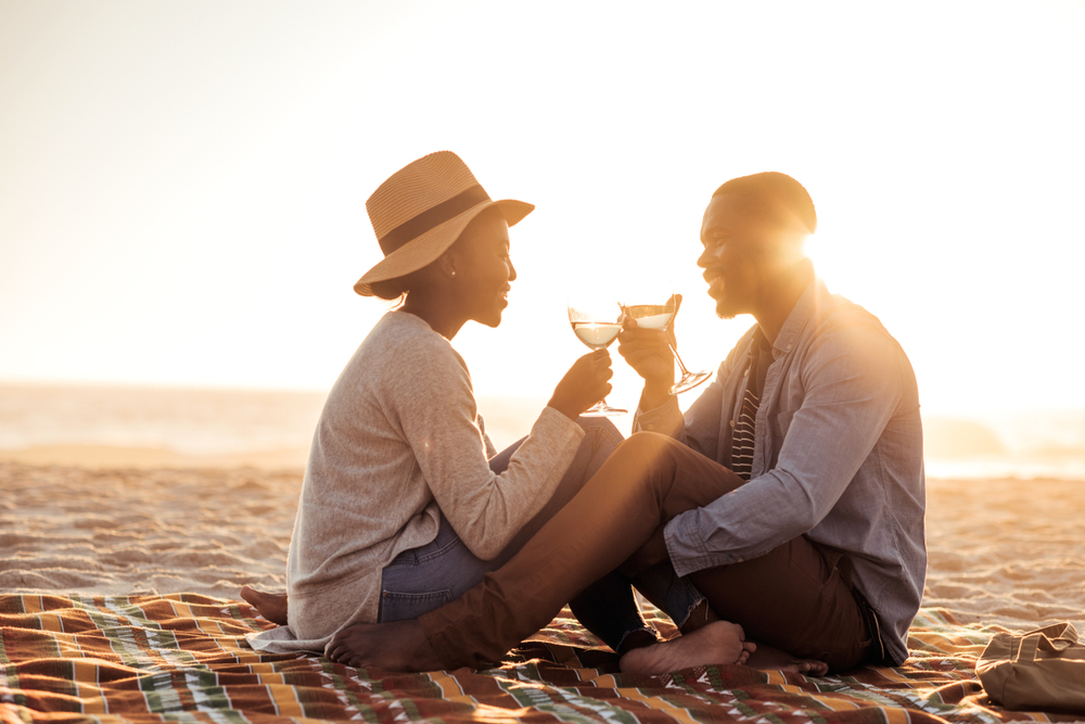 Romantic young African couple toasting each other with wine while sitting together on a sandy beach at dusk