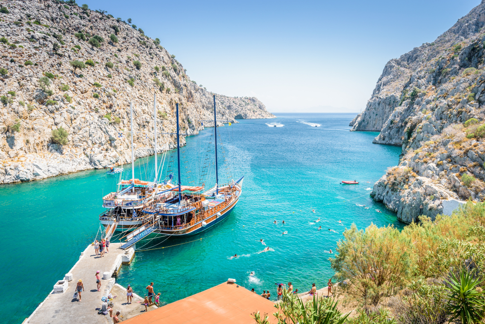 Beautiful sunny coast view to the greek blue sea with crystal clear water beach with big tourist boat cruising around pier in Vathys Bay