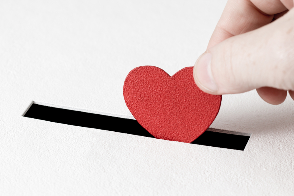 Red heart symbol is put by person's hand into slot of white donation box. Concept of sincere devotion to faith. Concept of donorship, life saving or charity. Close-up shot