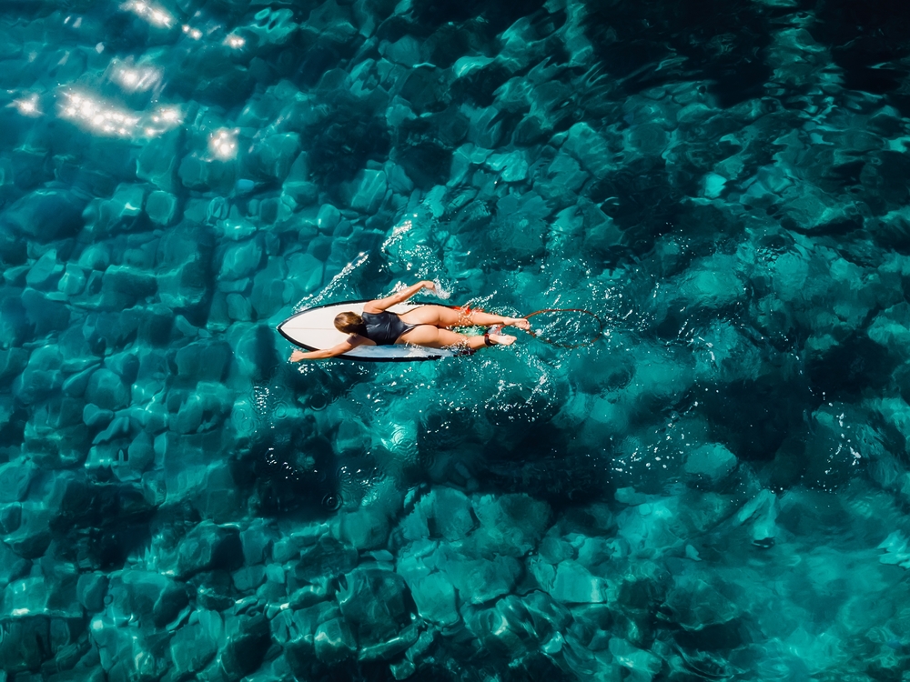 Surfer woman rowing on surfboard in turquoise ocean. Aerial drone view with surfgirl