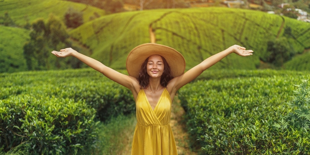 Romantic stylish woman traveler standing with raised hands against nature background tea plantations landscape in Sri Lanka. Portrait of young adult brunette woman in straw hat and yellow dress in