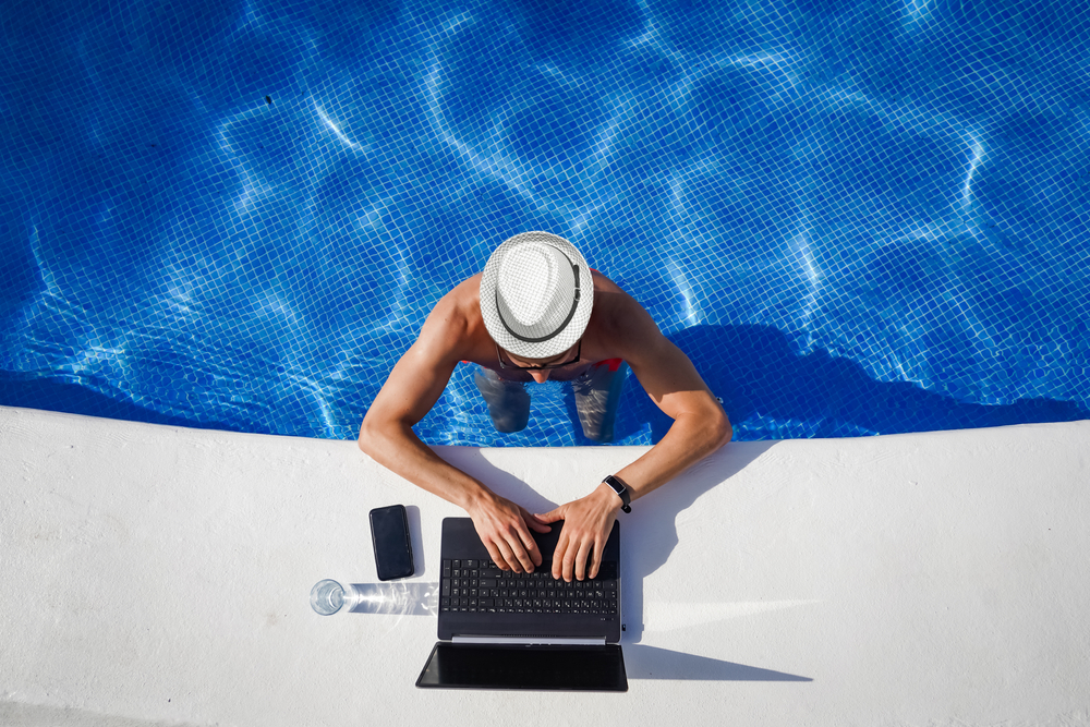 bird view of remote online working digital nomad man on workation with hat and laptop at a white table standing in a sunny turquoise water pool