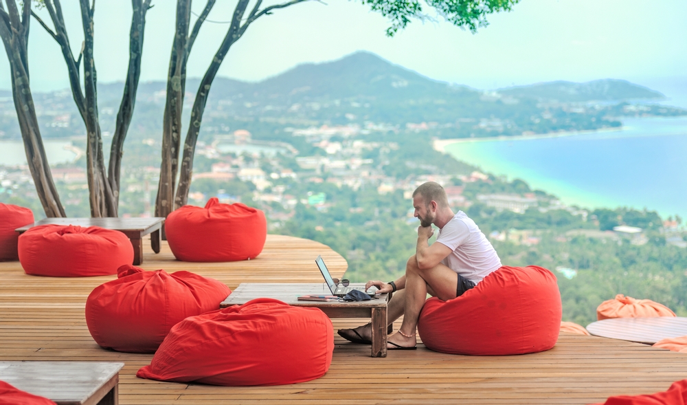 A Caucasian man sitting on a red cushion working remotely with his laptop.