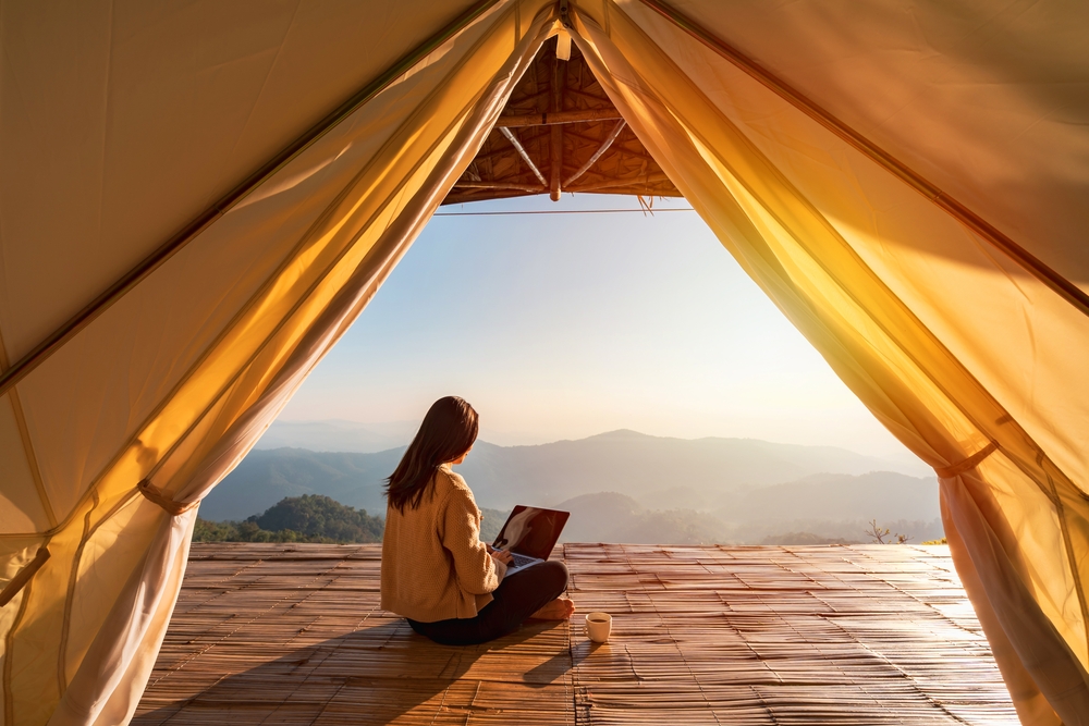 Young woman freelancer traveler working online using laptop and enjoying the beautiful nature landscape with mountain view at sunrise