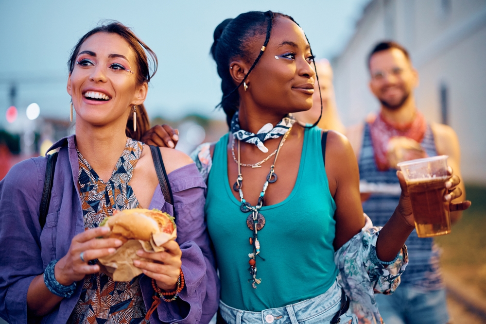 Happy female festival goers eating hamburger and drinking beer during open air music concert in summer.