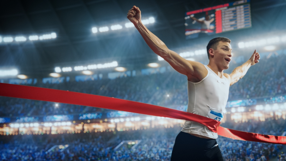 Professional Runner Competing In National Championship. A strong Caucasian Male Athlete is Sprinting Towards the Line in a Crowded Stadium, Crossing the Race Finish with His Hands in the Air