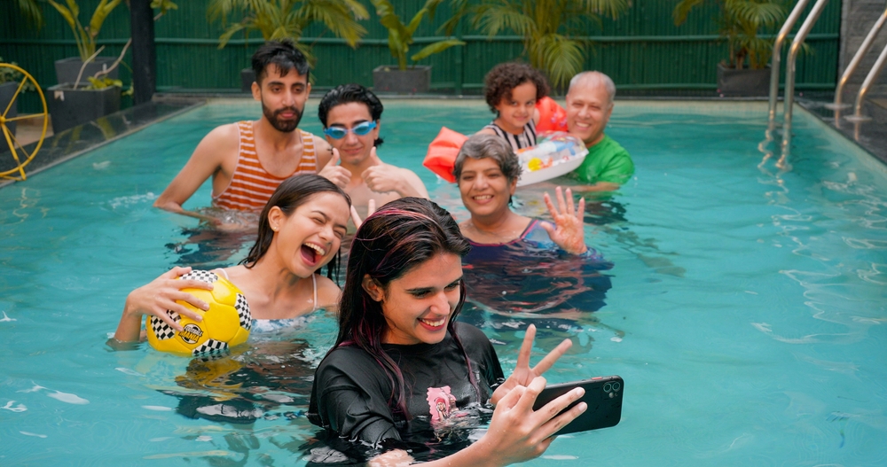 Happy Indian family having fun and enjoying outdoor picnic. Young girl taking selfie with senior parents using mobile smartphone at water park. Cute Child relax in colorful toy floating ring at resort