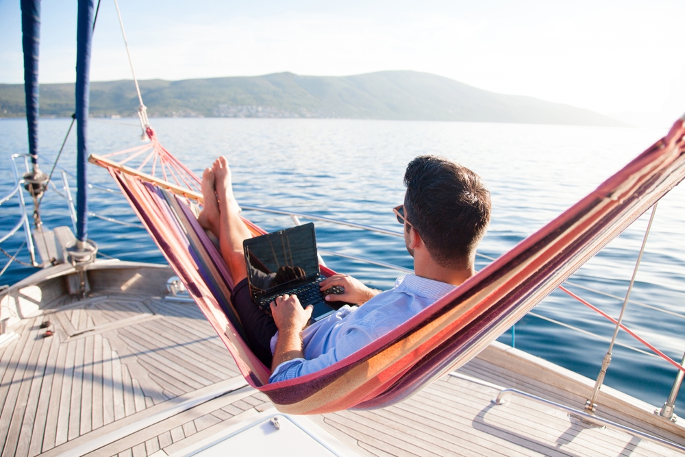 Working in sea traveling. Man with laptop in beach hammock. Summer vacation, living on yacht. Successful freelancer using computer. Workplace on nature outdoors, home office. Business lifestyle