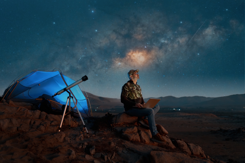 digital nomad man sitting alone with a laptop in the desert at night next to a telescope watching the starry night