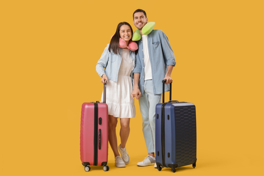 Beautiful young happy couple of tourists with suitcases and travel pillows on yellow background