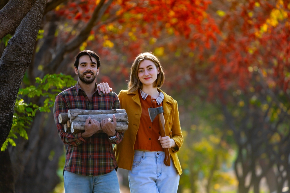 Happy caucasian farmer couple carrying chopped firewood while walking along countryside with fall color from maple tree during autumn season for winter preparation and off-grid living in rural home