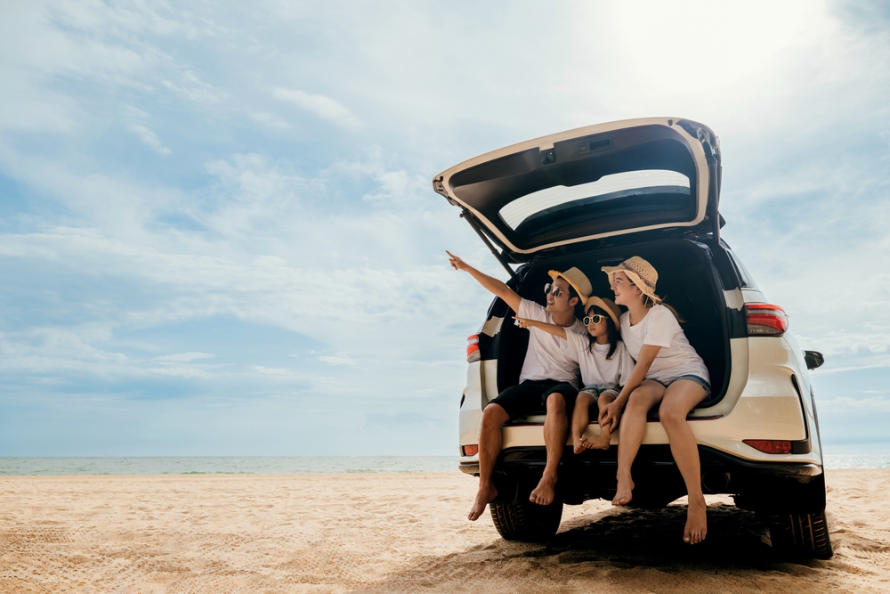 Dad, mom and daughter enjoying road trip sitting on back car and pointing finger out blue sky