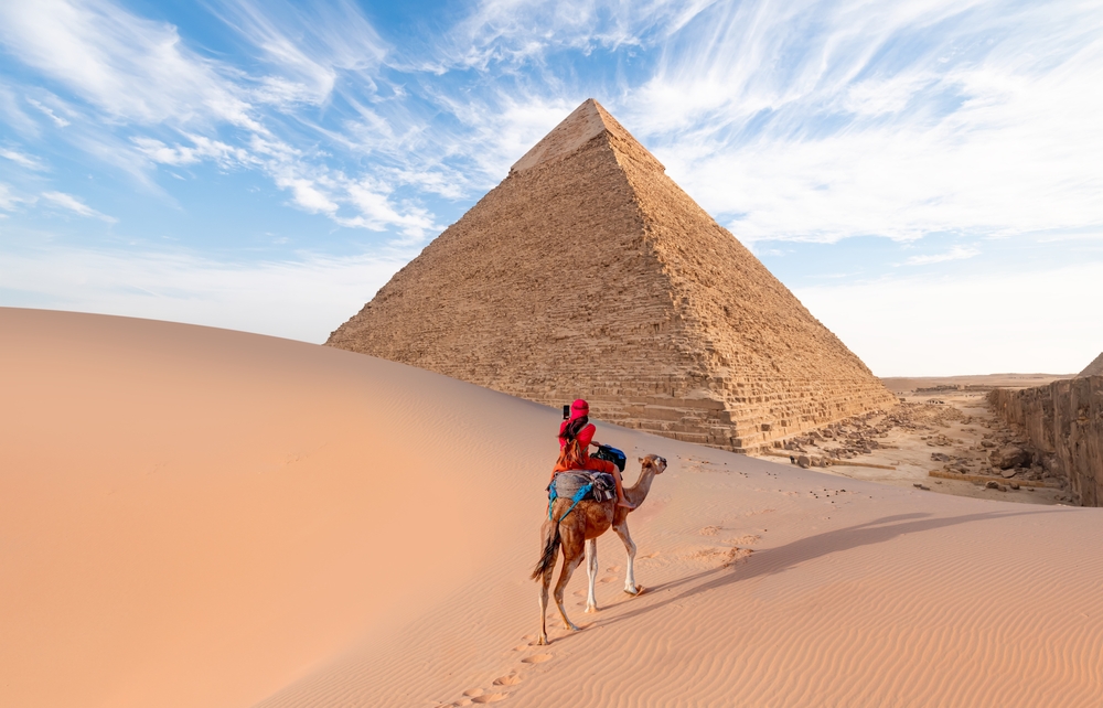 Camels in Giza Pyramid Complex - A woman in a red turban riding a camel across the thin sand dunes - Cairo, Egypt