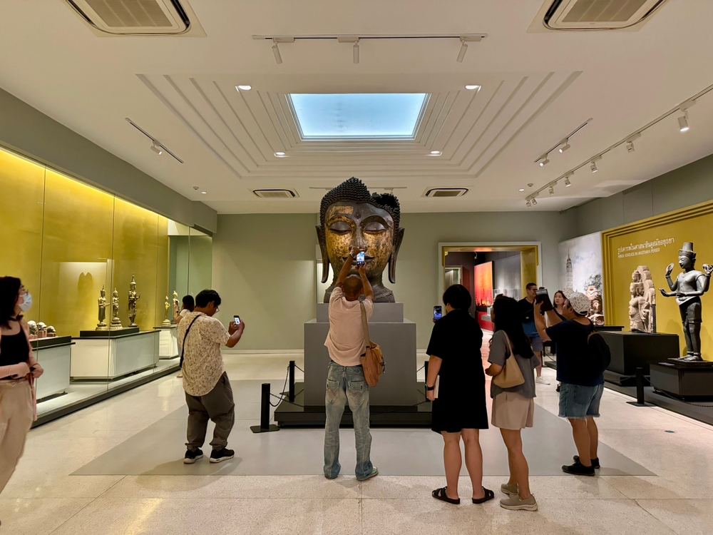 People stand and take photos of the Buddha head from the Ayutthaya period in the middle of the exhibition room, The National Museum Bangkok.