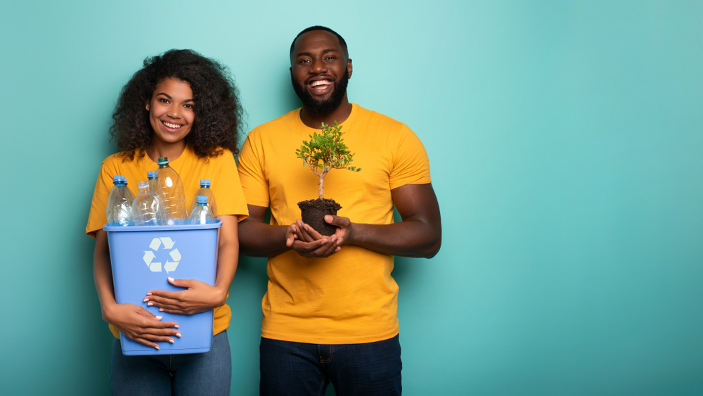 Happy couple hold a plastic container and a small tree over a light blue color.