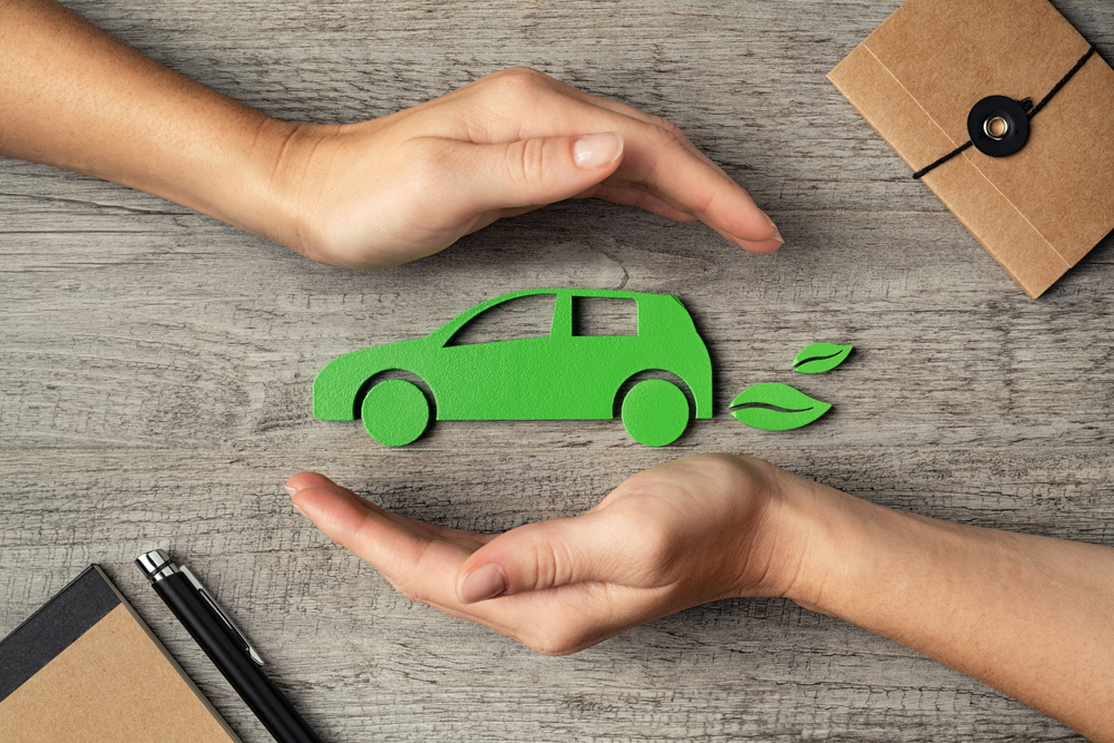 Close up of hands of woman protecting icon of green eco-friendly car over wooden table. 