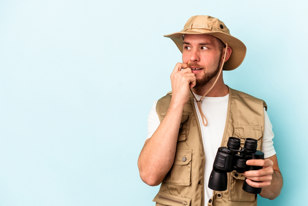 Young caucasian man looking at animals through binoculars isolated on blue background relaxed thinking about something looking at a copy space.