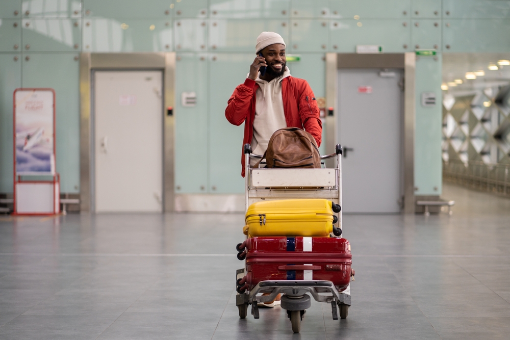 Young smiling African American man pushing luggage trolley while walking after arrival at airport, talking on mobile phone.