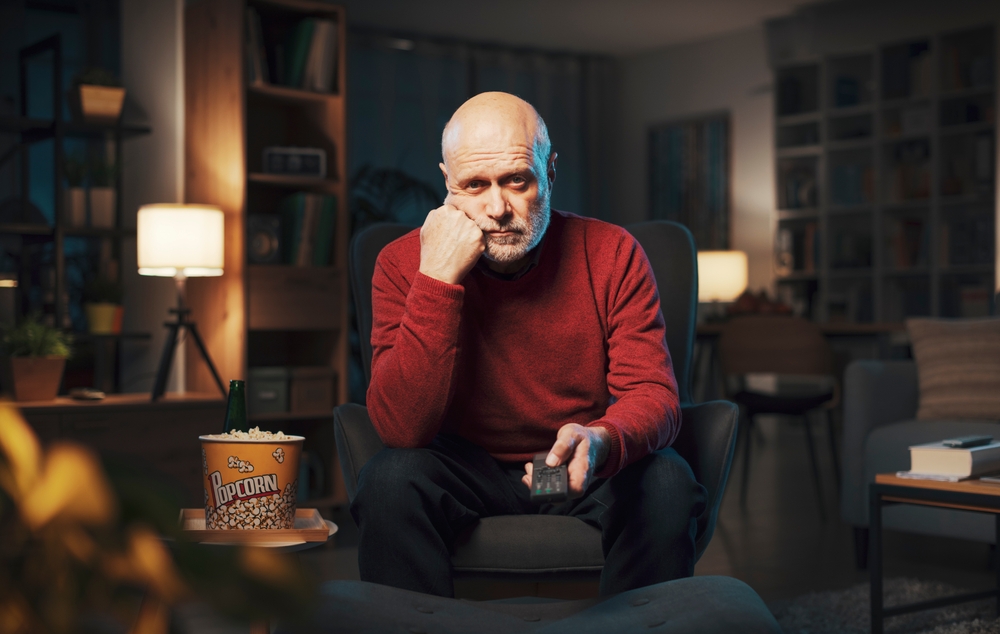 Bored senior man sitting on the armchair and watching TV, he is holding the remote control and changing channels