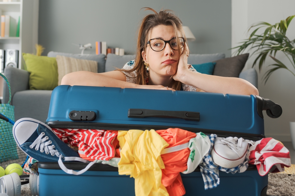 Tired sad woman packing for a long journey, she is leaning on her overfilled trolley bag