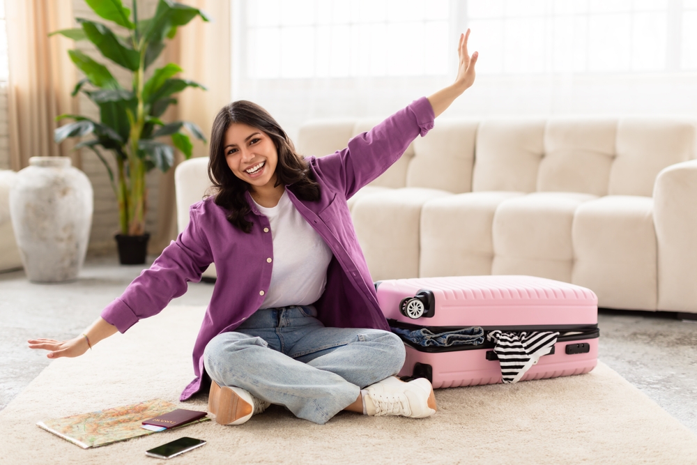 A joyous middle eastern woman with arms wide open sitting on the floor next to a pink suitcase, expressing travel excitement