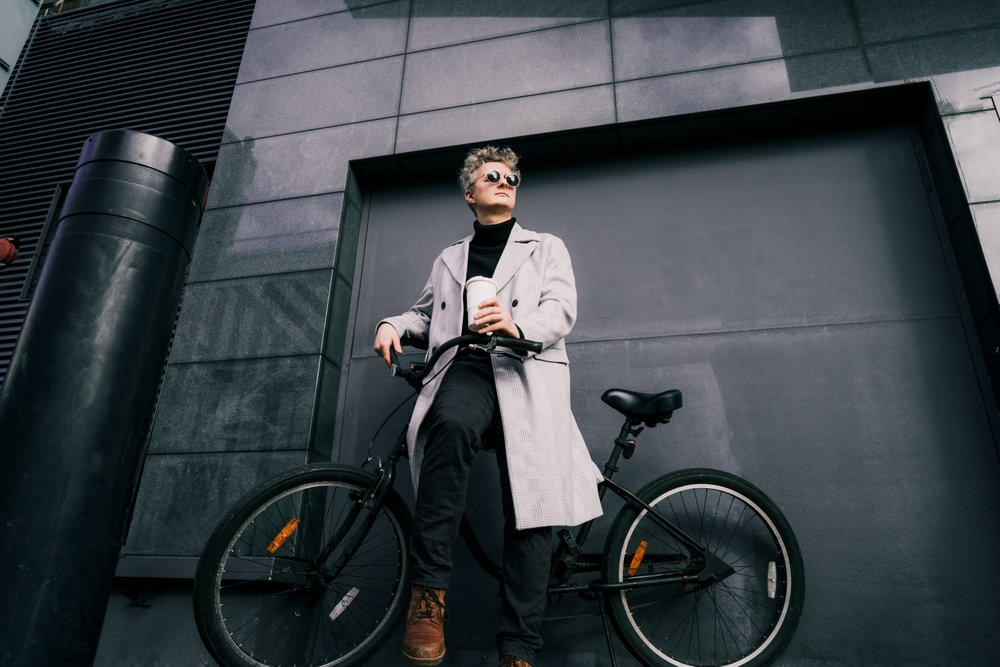 Stylish male in coat and sunglasses enjoying coffee out of reusable cup while sitting on retro bicycle near gray urban wall.