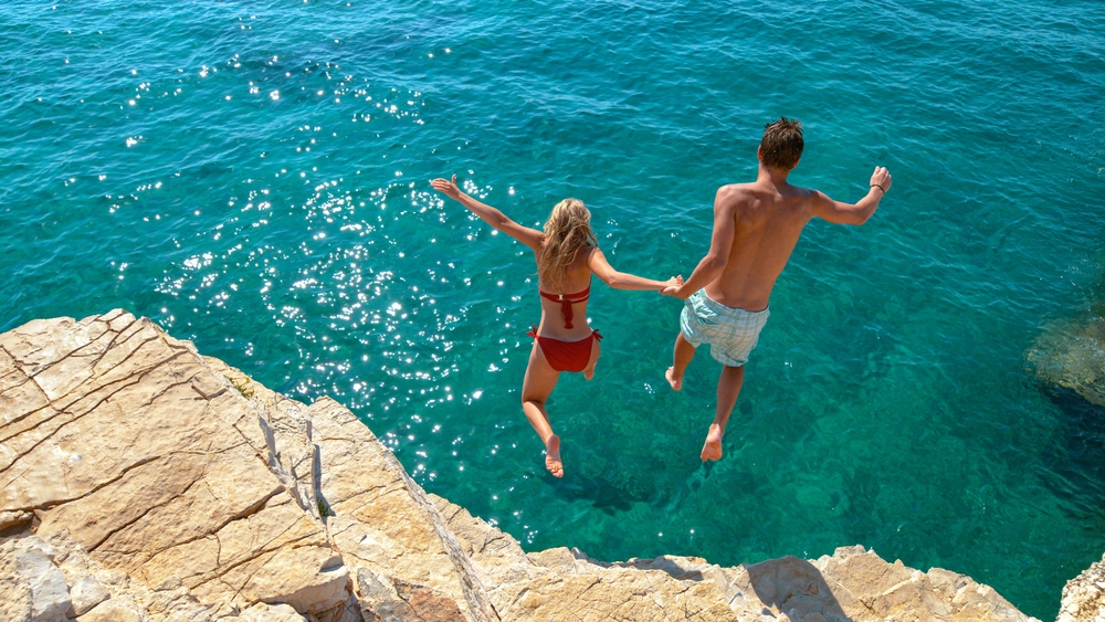 Carefree tourists hold hands while jumping into the refreshing blue sea during a relaxing summer vacation.