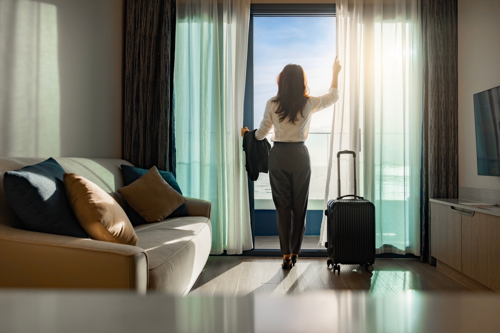 Confident businesswoman wearing white shirt and holding suit with suitcase stands in a hotel room while looking at the view from window Beautiful sunset.